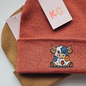 May include: Rust-colored knit beanie with a folded brim, featuring an embroidered cartoon cow with patchwork details. A second, tan beanie is visible beneath. A tag with the text "KC studios" is attached to the rust-colored beanie.
