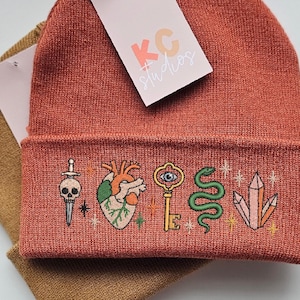 May include: Two beanies, one rust-colored and one tan, are stacked. The rust-colored beanie has a folded brim embroidered with a skull, heart, key, snake, and crystals. A tag reads "KC studios".