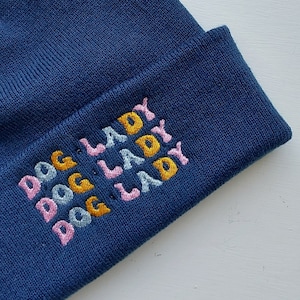 May include: A blue knitted beanie with the text "DOG LADY" embroidered in pink, light blue, and yellow.