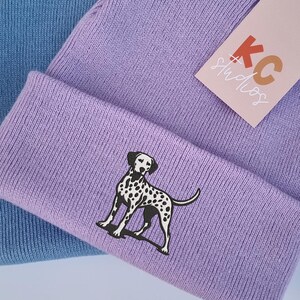 May include: Two knitted beanies, one light blue and one lavender, with a black and white dalmatian dog graphic on the lavender beanie. The lavender beanie has a tag that says "KC Studios".