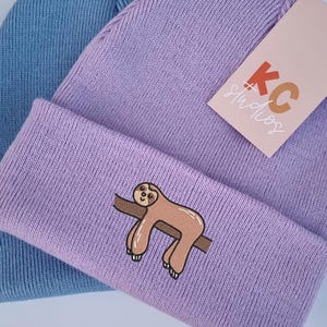 Può includere: Due berretti in maglia. Uno è azzurro, l'altro è lavanda con un bordo ripiegato. Il berretto lavanda ha una grafica di bradipo marrone sul bordo. Un'etichetta con la scritta "KC Studios" è attaccata al berretto lavanda.