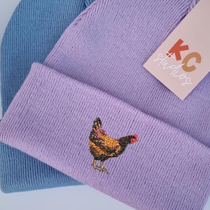 May include: Two knit beanies, one light blue and one lavender. The lavender beanie features an embroidered chicken design and a tag that reads "KC Studios". The beanies are folded over.