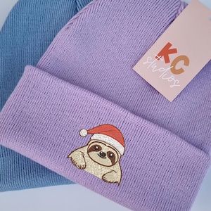 Può includere: Due berretti in maglia, uno azzurro chiaro e uno lavanda. Il berretto lavanda presenta un bradipo ricamato con un cappello da Babbo Natale. Un'etichetta con il testo "KC Studios" è attaccata al berretto lavanda.