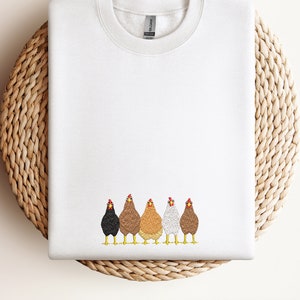 Può includere: Felpa bianca con una fila di cinque galline ricamate in diversi colori. Le galline sono rivolte in avanti con la testa alta.