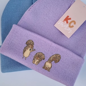 May include: Two knitted beanies, one light blue and one lavender, with a tag that says "KC Studios" on the lavender beanie. The lavender beanie has an embroidered design of three squirrels.