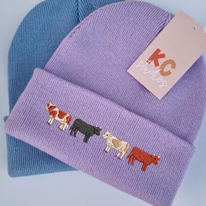 Op de afbeelding: Twee gebreide mutsen. E&eacute;n is lichtblauw, de andere is lavendelkleurig met een geborduurd ontwerp van vier koeien in verschillende kleuren. Een label van KC Studios is aan de lavendelkleurige muts bevestigd.