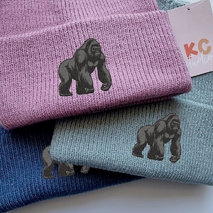 May include: Three knitted beanies in different colours, pink, blue and green, with a black gorilla graphic embroidered on each. The beanies are stacked on top of each other. The pink beanie has a tag that says "KC Studios".