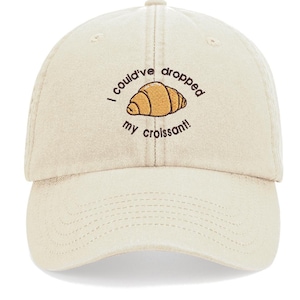 May include: A beige baseball cap with the text "I could've dropped my croissant!" embroidered on the front. The text is in brown and black, and there is a small embroidered image of a croissant.