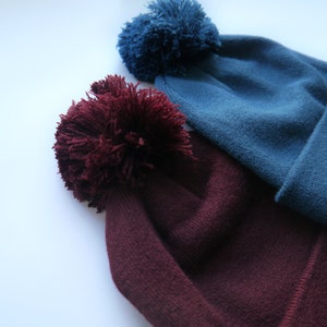 Op de afbeelding: Twee gebreide mutsen met grote pompons. De ene muts is diep bordeauxrood, de andere is blauwgroen. De mutsen zijn gemaakt van zacht, getextureerd breiwerk. De pluizige pompons geven een speelse uitstraling.