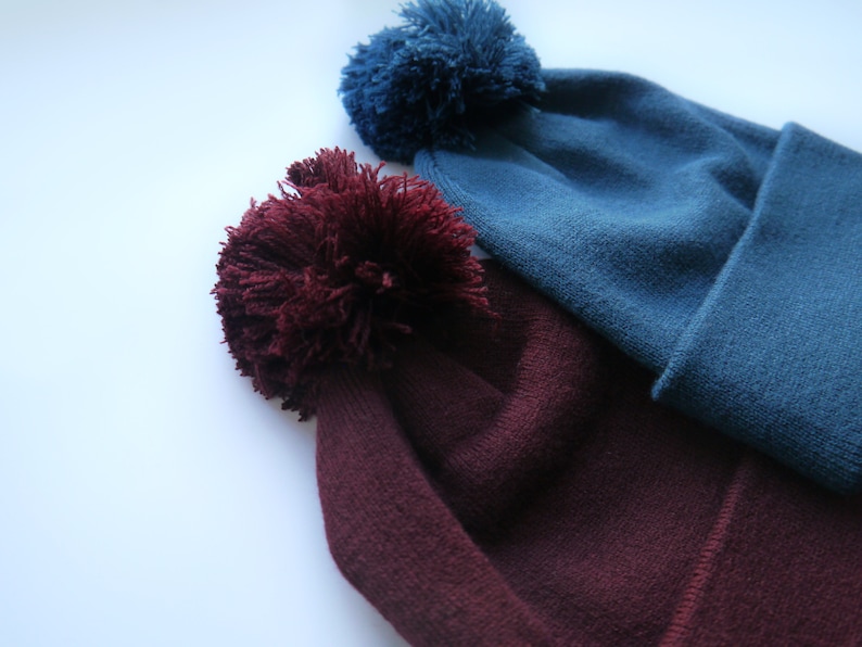 Op de afbeelding: Twee gebreide mutsen met grote pompons. De ene muts is diep bordeauxrood, de andere is blauwgroen. De mutsen zijn gemaakt van zacht, getextureerd breiwerk. De pluizige pompons geven een speelse uitstraling.