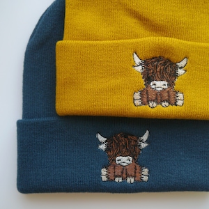 K&ouml;nnte beinhalten: Zwei Strickm&uuml;tzen mit einem gestickten Highland-Cow-Motiv. Die obere M&uuml;tze ist senfgelb und die untere M&uuml;tze ist dunkelblau.