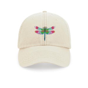 Könnte beinhalten: Cremefarbene Baseballkappe mit einem farbenfrohen Libellen-Design. Die Libelle hat rosa, blaue, grüne und violette Flügel und einen blauen Körper. Die Kappe ist aus weichem Stoff und hat einen gebogenen Schirm.