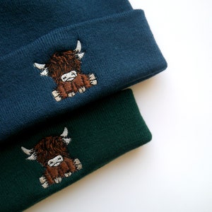 K&ouml;nnte beinhalten: Zwei Strickm&uuml;tzen mit einer braunen Highland-Kuh, die auf der Vorderseite gestickt ist. Eine M&uuml;tze ist dunkelblau und die andere dunkelgr&uuml;n.