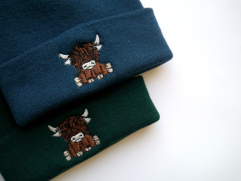 K&ouml;nnte beinhalten: Zwei Strickm&uuml;tzen mit einer braunen Highland-Kuh, die auf der Vorderseite gestickt ist. Eine M&uuml;tze ist dunkelblau und die andere dunkelgr&uuml;n.