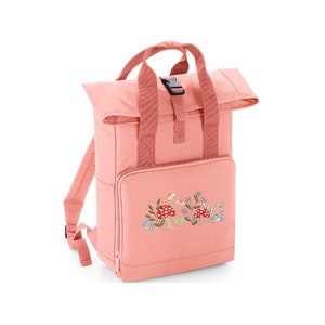 May include: A pink canvas backpack with a roll-top closure and a front pocket. The backpack has an embroidered design of mushrooms and flowers.