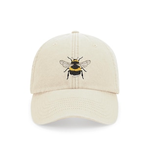 Könnte beinhalten: Eine beige Baseballkappe mit einem gestickten Hummel-Design. Die Hummel ist schwarz und gelb mit braunen Flügeln.