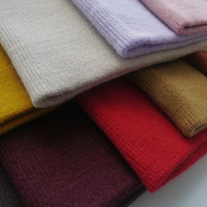 Op de afbeelding: Close-up van een stapel gevouwen gebreide mutsen in verschillende kleuren. De kleuren zijn onder meer mosterdgeel, lichtgrijs, lavendel en rood. De mutsen zijn gemaakt van een zacht, getextureerd breimateriaal.