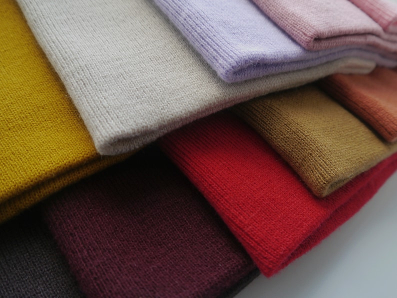 Op de afbeelding: Close-up van een stapel gevouwen gebreide mutsen in verschillende kleuren. De kleuren zijn onder meer mosterdgeel, lichtgrijs, lavendel en rood. De mutsen zijn gemaakt van een zacht, getextureerd breimateriaal.