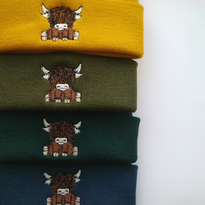 K&ouml;nnte beinhalten: Vier gestrickte Beanies in verschiedenen Farben, jede mit einem gestickten Highland-Kuh-Design. Die Beanies sind &uuml;bereinander gestapelt, beginnend mit einem gelben Beanie, gefolgt von einem gr&uuml;nen Beanie, einem dunkelgr&uuml;nen Beanie und einem blauen Beanie.