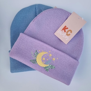 Può includere: Due berretti lavorati a maglia nell'immagine. Uno è azzurro, l'altro è lavanda con ricami dorati a forma di luna crescente e stelle. Un'etichetta con la scritta "KC Studios" è attaccata al berretto lavanda.