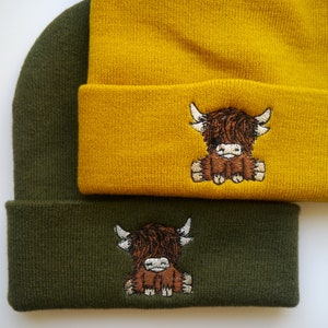 K&ouml;nnte beinhalten: Zwei Strickm&uuml;tzen mit einem Highland-Cow-Design. Eine M&uuml;tze ist senfgelb und die andere ist waldgr&uuml;n.