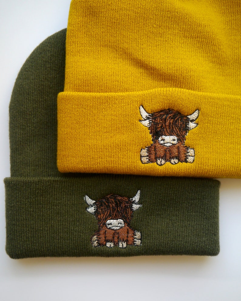 K&ouml;nnte beinhalten: Zwei Strickm&uuml;tzen mit einem Highland-Cow-Design. Eine M&uuml;tze ist senfgelb und die andere ist waldgr&uuml;n.