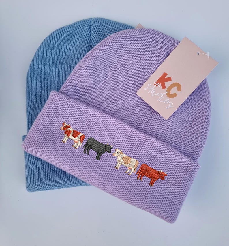 Op de afbeelding: Twee gebreide mutsen. E&eacute;n is lichtblauw, de andere is lavendelkleurig met een geborduurd ontwerp van vier koeien in verschillende kleuren. Een label van KC Studios is aan de lavendelkleurige muts bevestigd.
