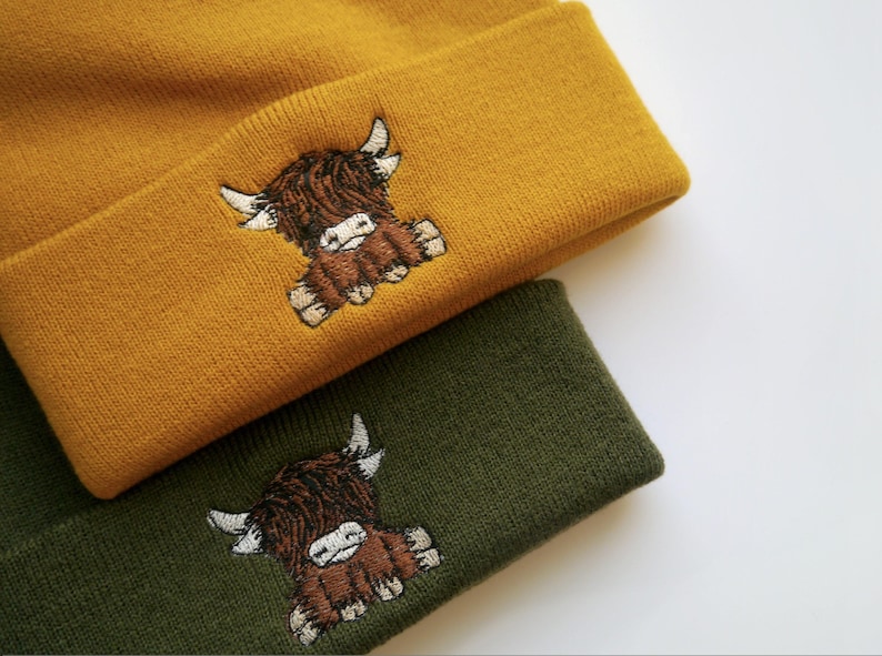 K&ouml;nnte beinhalten: Zwei Strickm&uuml;tzen mit gestickten Hochlandrind-Motiven. Eine M&uuml;tze ist senfgelb und die andere ist waldgr&uuml;n.