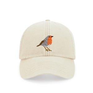 以下が含まれることがあります： 前面に刺繍されたコマドリがついたベージュのベースボールキャップ。コマドリは灰色と白で、胸が赤いです。