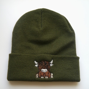 K&ouml;nnte beinhalten: Eine gr&uuml;ne Strickm&uuml;tze mit einem gestickten Highland-Rind in Braun und Wei&szlig;.