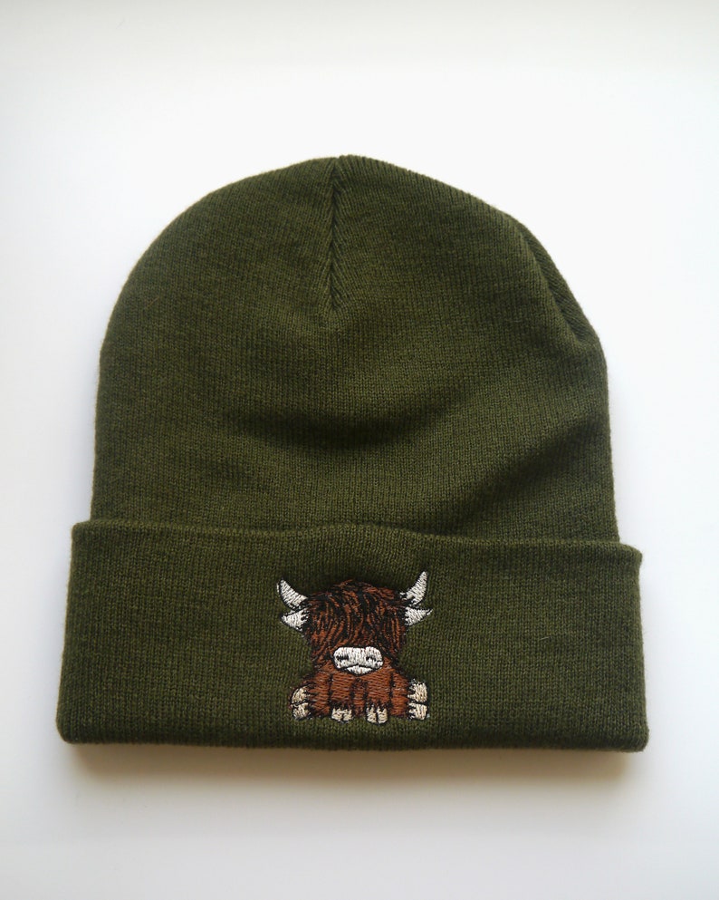 K&ouml;nnte beinhalten: Eine gr&uuml;ne Strickm&uuml;tze mit einem gestickten Highland-Rind in Braun und Wei&szlig;.