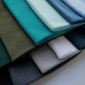 Op de afbeelding: Een verzameling gevouwen gebreide stoffen in verschillende tinten blauw, groen, grijs en wit. De kleuren vari&euml;ren van diep teal en bosgroen tot lichtblauw en grijs. De stof lijkt zacht en heeft een geribbelde textuur.