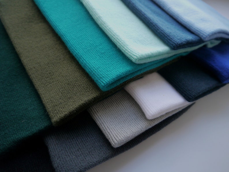 Op de afbeelding: Een verzameling gevouwen gebreide stoffen in verschillende tinten blauw, groen, grijs en wit. De kleuren vari&euml;ren van diep teal en bosgroen tot lichtblauw en grijs. De stof lijkt zacht en heeft een geribbelde textuur.