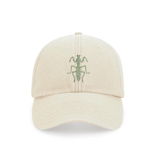 Op de afbeelding: Een beige baseballpet met een geborduurde groene bidsprinkhaan op de voorkant.