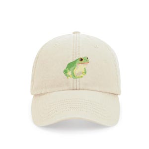 May include: A beige baseball cap featuring an embroidered green frog with red eyes. The frog is positioned on the front of the cap, which has a curved brim and an adjustable strap at the back. The cap is made of a soft, durable fabric.