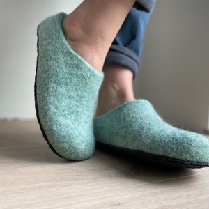 Könnte beinhalten: Ein Paar hellgrüne Wollhausschuhe mit schwarzen Sohlen. Die Hausschuhe sind aus einem weichen, flauschigen Material gefertigt und haben eine bequeme Passform.
