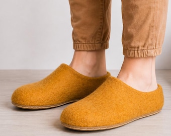May include: A pair of mustard yellow felted wool slippers with a rounded toe and a flat sole. The slippers feature a simple slip-on design with a seamless construction, providing a clean look. The material has a textured, soft appearance.