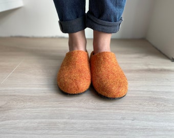 May include: A pair of orange felt slippers with a black trim. The slippers are being worn by someone standing on a light wood floor.