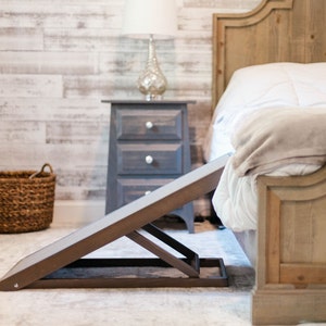 May include: A brown wooden pet ramp with a textured surface. The ramp is folded up and is in front of a wooden bed frame with a white bedspread and a tan blanket.