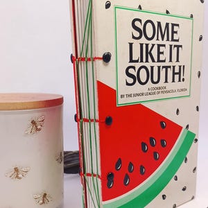 May include: A cookbook titled "Some Like It South!" with a watermelon slice illustration. The book is bound with red and green thread and black fasteners. A candle with bee designs and a pen are also visible.