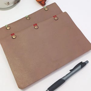 May include: Two brown leather-like menu covers with brass rivets and colored ribbon accents. A black and silver pen rests on a white surface. Decorative flowers and a glass vase are in the background.