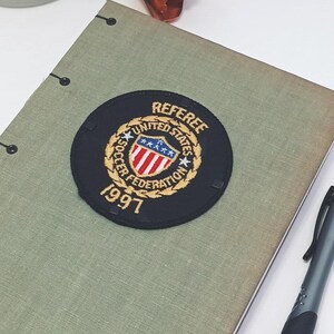 May include: A light green notebook with a black and gold patch that reads "REFEREE UNITED STATES SOCCER FEDERATION 1997". A black and silver pen rests on the notebook. Decorative flowers and a white mug with bee designs are in the background.
