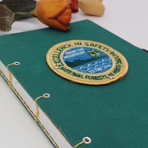 May include: A green journal with a sewn binding and a patch that reads "Excellence in Safety". The patch features a mountain and water scene. A decorative flower is in the background.