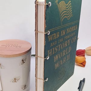 May include: A vintage book titled "War in Korea and the Complete History of World War II" with a green cover and gold lettering. The book is bound with visible stitching and metal fasteners. A candle and pen are also in the image.