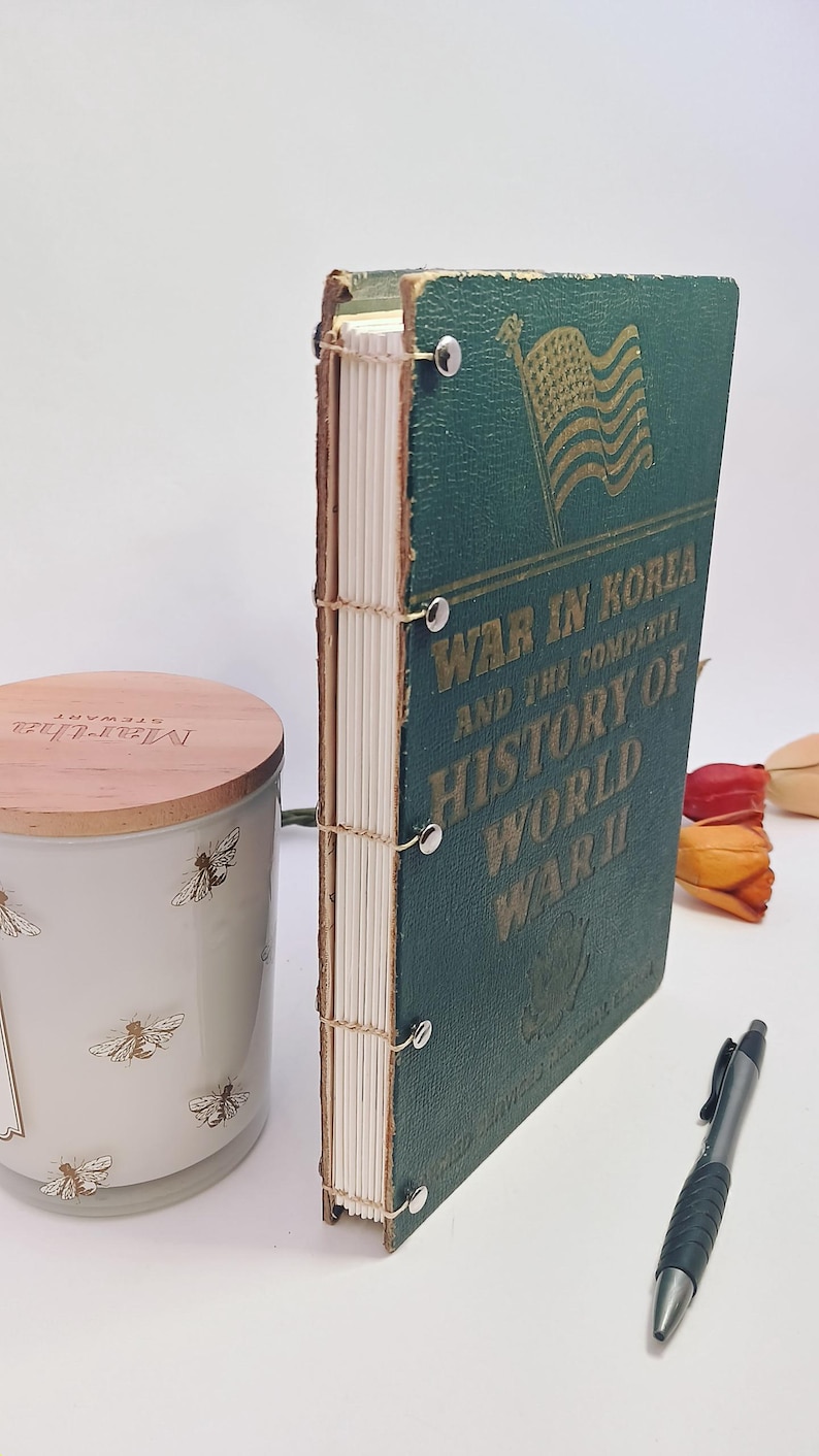 May include: A vintage book titled "War in Korea and the Complete History of World War II" with a green cover and gold lettering. The book is bound with visible stitching and metal fasteners. A candle and pen are also in the image.