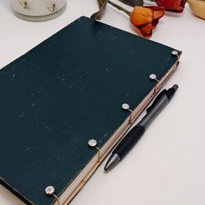 May include: A dark teal journal with silver binding posts and a black pen. A white jar with gold bee designs and faux flowers are in the background. The journal is closed and has a textured cover.