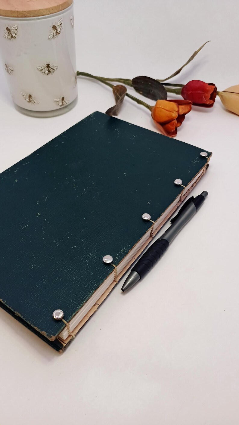 May include: A dark teal journal with silver binding posts and a black pen. A white jar with gold bee designs and faux flowers are in the background. The journal is closed and has a textured cover.