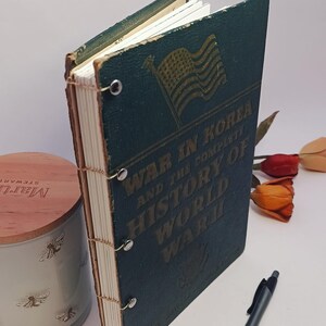 May include: A vintage book titled "War in Korea and the Complete History of World War II" with an American flag graphic. The book has a dark teal cover and is bound with metal rings. A pen and a decorative candle are also in the image.