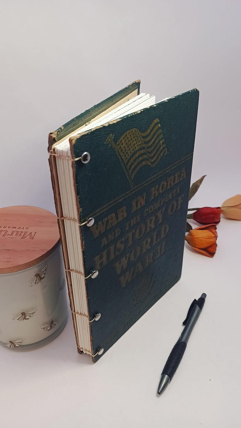 May include: A vintage book titled "War in Korea and the Complete History of World War II" with an American flag graphic. The book has a dark teal cover and is bound with metal rings. A pen and a decorative candle are also in the image.