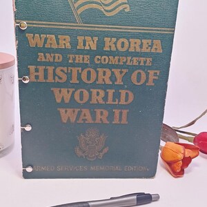 May include: A vintage book titled "War in Korea and the Complete History of World War II." The cover is dark green with gold lettering and an illustration of the American flag. A silver and black pen rests in front of the book.